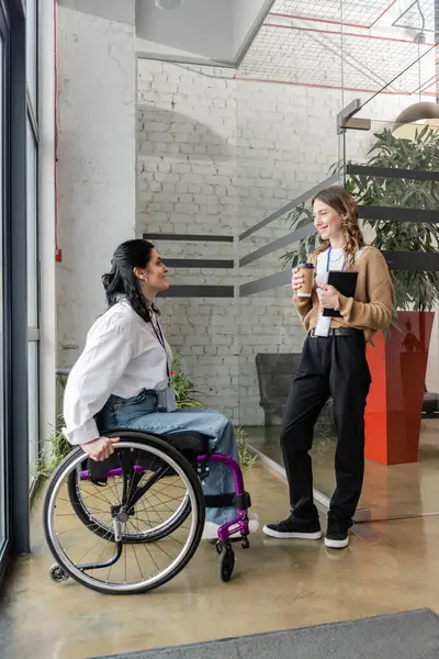 Концепция включения, счастливая женщина, держащая кофе и беседующая с коллегой-инвалидом у стеклянной двери — стоковое фото