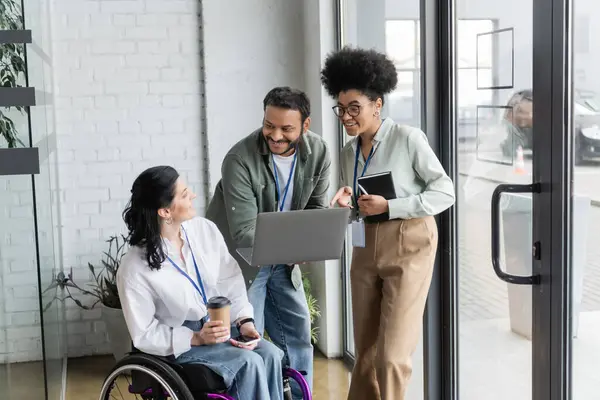 Happy disabled woman on wheelchair looking at indian man with laptop, diverse business people — Stock Photo