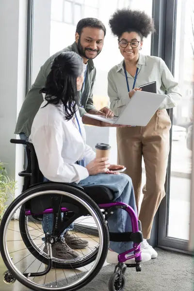 Heureux collègues interraciaux regardant collègue handicapé en fauteuil roulant, discuter de la stratégie de travail — Photo de stock