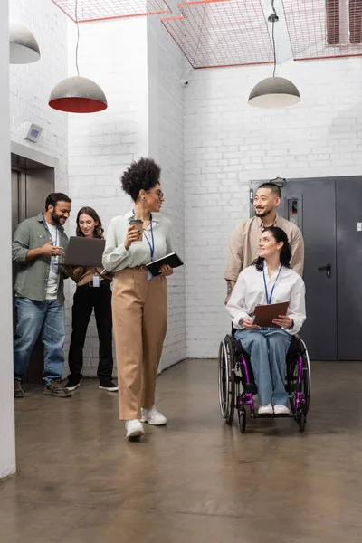 Homme asiatique poussant fauteuil roulant de femme d'affaires handicapée et regardant un collègue afro-américain — Photo de stock