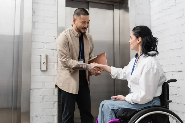 Включение и разнообразие, веселый азиатский мужчина пожимающий руку женщине-инвалиду возле офисных лифтов — стоковое фото