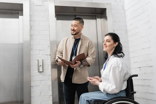 Включение и разнообразие, счастливый азиатский мужчина папку рядом с инвалидом женщина и офисные лифты, смотреть в сторону — стоковое фото