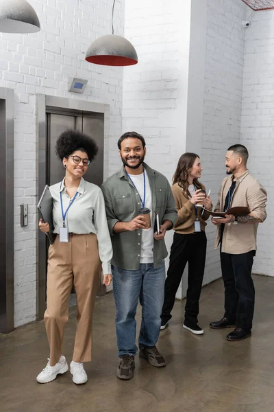 Heureuse femme afro-américaine et homme indien regardant la caméra près des ascenseurs de bureau, diverses personnes — Photo de stock