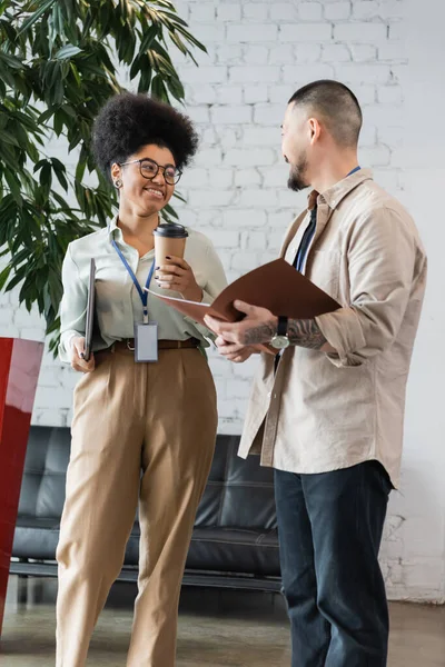 Cheerful african american woman holding coffee to go and chatting with asian coworker during break — Stock Photo