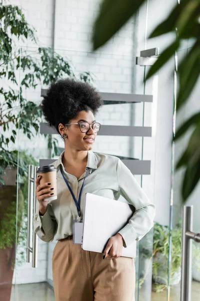 Fröhliche afrikanisch-amerikanische Geschäftsfrau in Gläsern mit Pappbecher und Laptop im modernen Büro — Stockfoto
