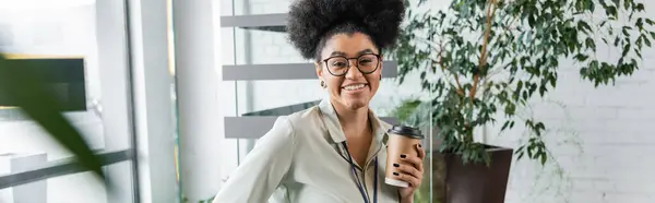 Happy african american businesswoman in glasses holding paper cup with coffee in office, banner — Stock Photo