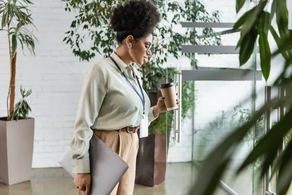 Vista lateral de la mujer de negocios afroamericana en gafas que sostienen el ordenador portátil y mirando la taza de papel - foto de stock