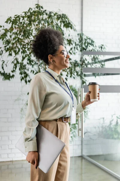 Vue latérale de heureuse femme d'affaires afro-américaine dans des lunettes tenant ordinateur portable et café à emporter — Photo de stock