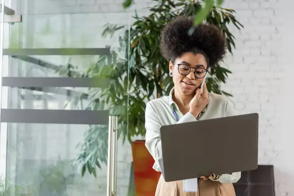 Femme d'affaires afro-américaine dans des lunettes à l'aide d'un ordinateur portable et parler sur smartphone, multitâche — Photo de stock