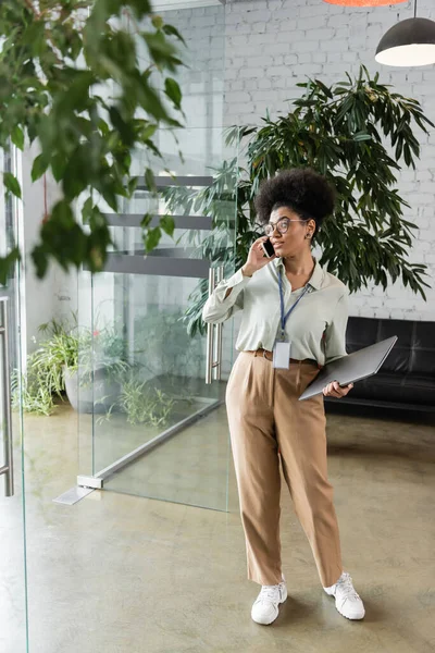 Mujer de negocios afroamericana en gafas que sostienen el ordenador portátil y hablan en el teléfono inteligente, longitud completa — Stock Photo