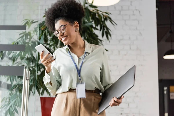 Позитивна афроамериканська бізнес-леді в окулярах тримає ноутбук і використовує смартфон в офісі — стокове фото