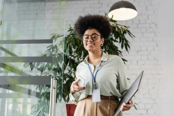 Портрет щасливої афроамериканської бізнес-леді в окулярах, що тримає ноутбук і смартфон в офісі — стокове фото