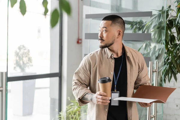 Distraído asiático hombre de negocios con insignia caminando en oficina con carpeta y papel taza en manos - foto de stock