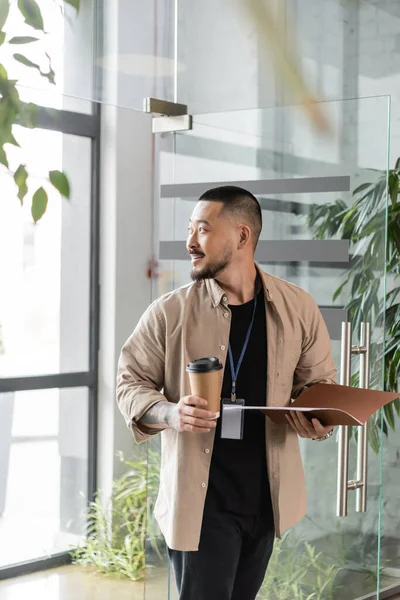 Distratto asiatico uomo d'affari con distintivo sorridente e camminando in ufficio con cartella e bicchiere di carta — Foto stock