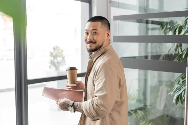 Heureux asiatique homme d'affaires avec tatouage souriant et marchant dans le bureau avec dossier et tasse en papier — Photo de stock