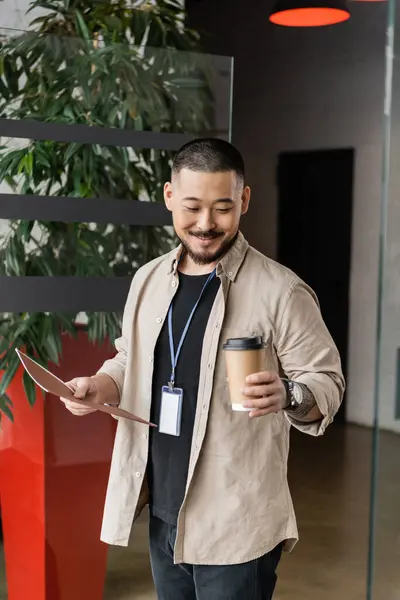 Alegre asiático hombre de negocios con tatuaje sonriendo y caminando en oficina con carpeta y papel taza - foto de stock