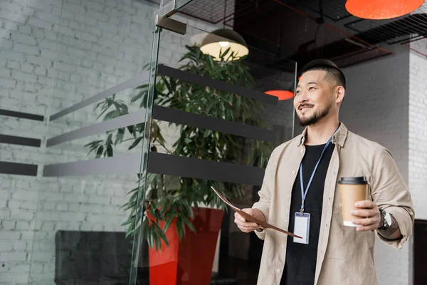 Alegre asiático hombre de negocios con tatuaje sonriendo y caminando en oficina con carpeta y café para ir - foto de stock
