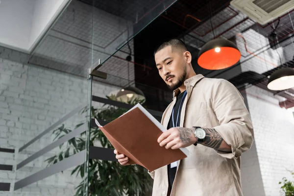Focused asian team leader with tattoo looking at startup plan inside of folder near glass door — Stock Photo
