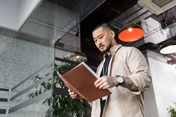 Concentré chef d'équipe asiatique avec tatouage regardant le plan de démarrage à l'intérieur du dossier près de la porte en verre — Photo de stock