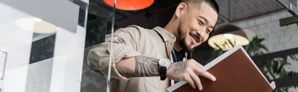 Feliz asiático hombre de negocios con tatuaje mirando documentos dentro de carpeta cerca de la puerta de vidrio, pancarta - foto de stock