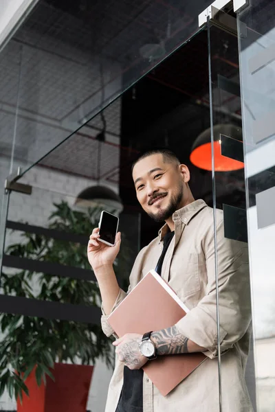 Feliz asiático hombre de negocios con tatuaje celebración carpeta y teléfono inteligente cerca de vidrio puerta de la oficina - foto de stock