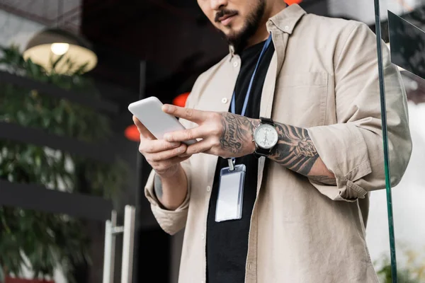 Vista recortada de hombre de negocios barbudo en ropa casual de pie con insignia y charlando en el teléfono inteligente - foto de stock