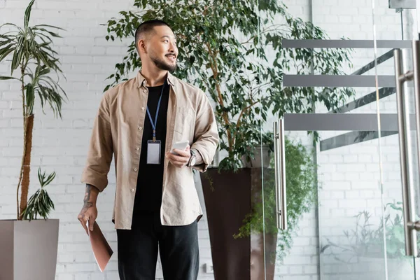 Joyful asian businessman with tattoo smiling while holding smartphone and folder in modern office — Stock Photo