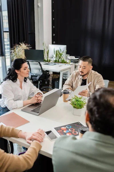 Asiático hombre mostrando gráficos a equipo líder sentado en trabajo escritorio cerca interracial colegas - foto de stock