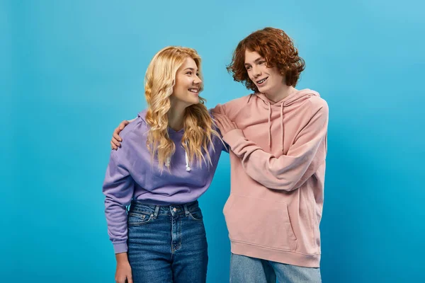 Feliz y elegante pareja de adolescentes abrazándose y sonriendo el uno al otro en azul, amistad y unidad - foto de stock