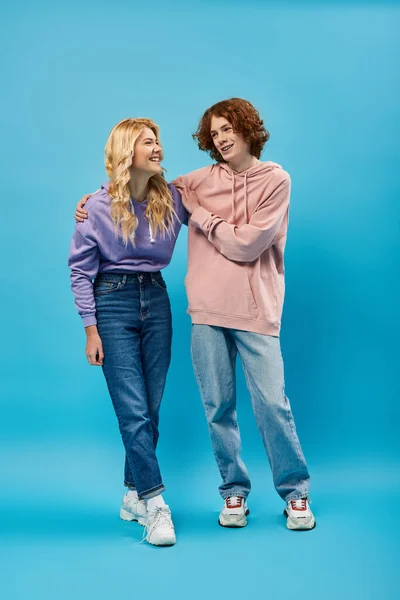 Allegri amici adolescenti con cappuccio e jeans che si abbracciano e si guardano l'un l'altro su blu, a tutta lunghezza — Foto stock