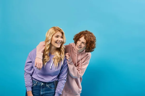 Rubia adolescente chica riendo cerca pelirroja novio, feliz amigos en elegante sudaderas con capucha y jeans en azul - foto de stock