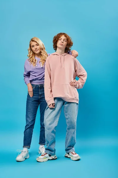 Fiducioso ragazzo adolescente rossa con mano sul fianco vicino sorridente ragazza bionda su blu, amici alla moda — Foto stock