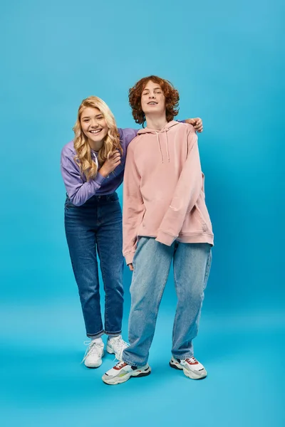 Alegre y elegante adolescente amigos en sudaderas con capucha y jeans sonriendo a la cámara en azul, longitud completa - foto de stock