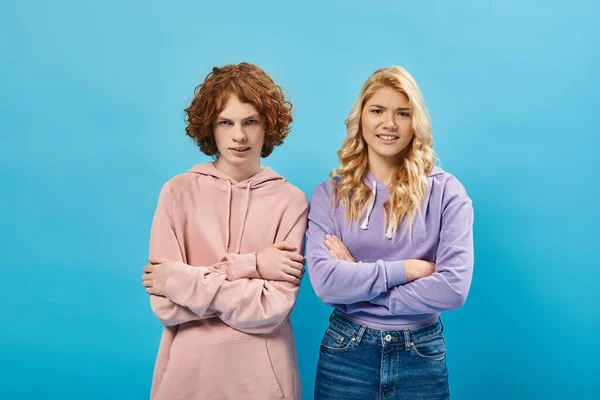 Irritated teenage friends in hoodies standing with crossed arms and looking at camera on blue — Stock Photo