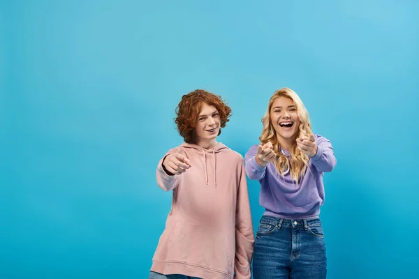 Cheerful and trendy teenage friends in hoodies pointing with fingers at camera and laughing on blue — Stock Photo