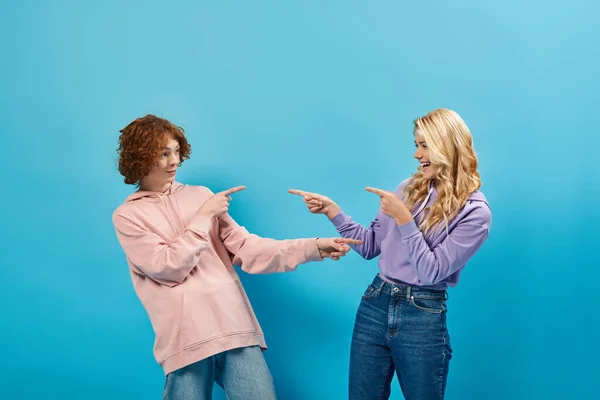 Joyful teenagers in stylish outfits pointing with fingers and each other and laughing on blue — Stock Photo