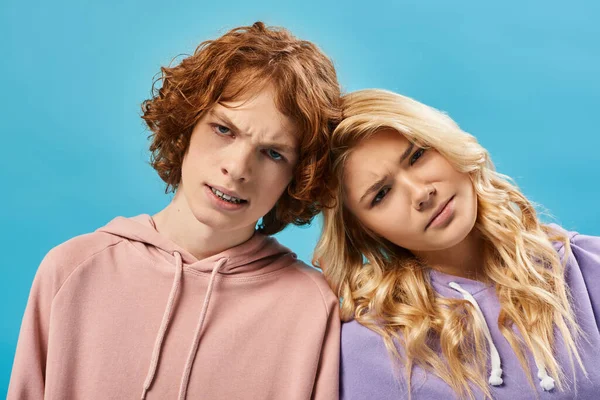 Portrait of frustrated teenagers, redhead boy and blonde girl in hoodies looking at camera on blue — Stock Photo