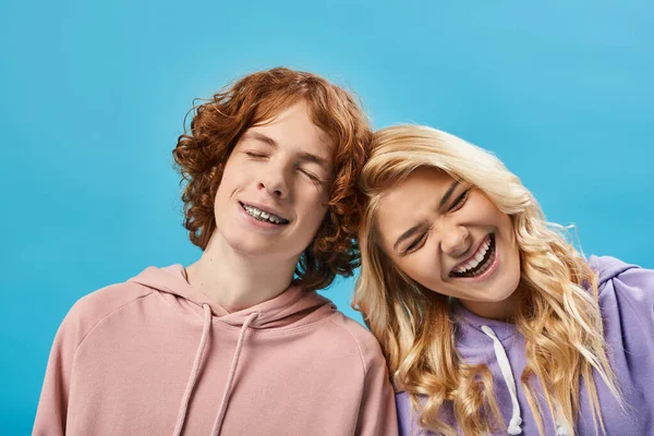 Portrait of excited teenage boy and girl in hoodies laughing with closed eyes on blue, happiness — Stock Photo