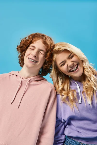 Amigos adolescentes llenos de alegría en sudaderas con capucha con estilo riendo con los ojos cerrados en azul, adolescentes despreocupados - foto de stock