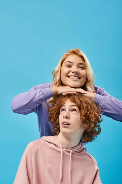 Expressive blonde teen girl having fun and smiling at camera above head of redhead boyfriend on blue — Stock Photo