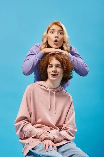 Amazed blonde teenage girl with open mouth looking at camera above head of redhead boyfriend on blue — Stock Photo