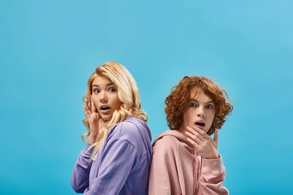 Shocked blonde teen girl with redhead boyfriend sitting back to back and looking at camera on blue — Stock Photo