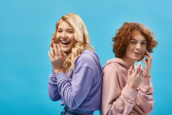 Excited teenage friends in hoodies sitting back to back and laughing at camera in studio on blue — Stock Photo