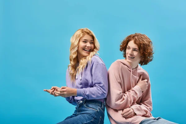 Amigos adolescentes felices en trajes de moda sentados espalda con espalda y sonriendo el uno al otro en azul - foto de stock