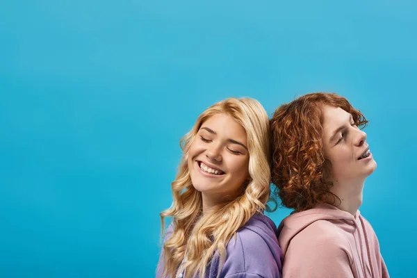 Positive teenage couple in stylish hoodies sitting back to back and smiling with closed eyes on blue — Stock Photo