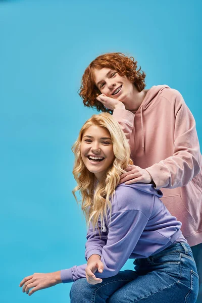 Счастливый рыжий подросток, опирающийся на блондинку, смотрящую в камеру и смеющийся над синим — стоковое фото