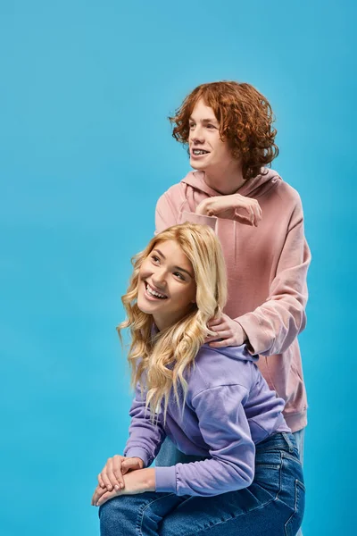 Cheerful redhead guy looking away near blonde girlfriend sitting on blue, happy teenage friends — Stock Photo