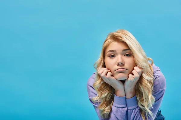 Frustrated blonde teenage girl in hoodie holding hands near face and looking at camera on blue — Stock Photo