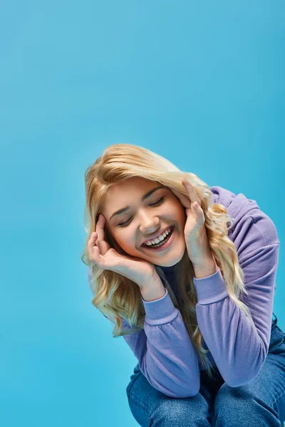 Cheerful blonde teen girl in hoodie laughing with hands near face and closed eyes on blue, happiness — Stock Photo