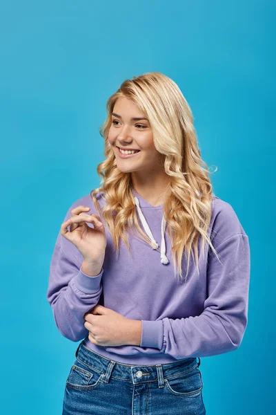 Позитивная блондинка-подросток в стильном капюшоне улыбается и смотрит в сторону в студии на синий — стоковое фото
